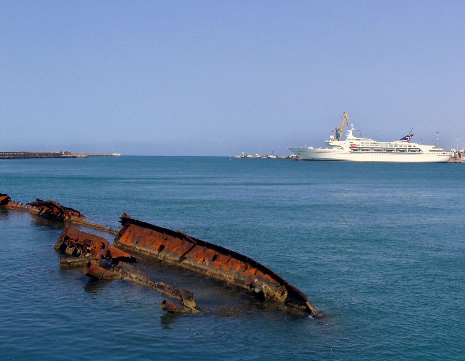 A rusted shipwreck in a harbour with a cruise ship behind