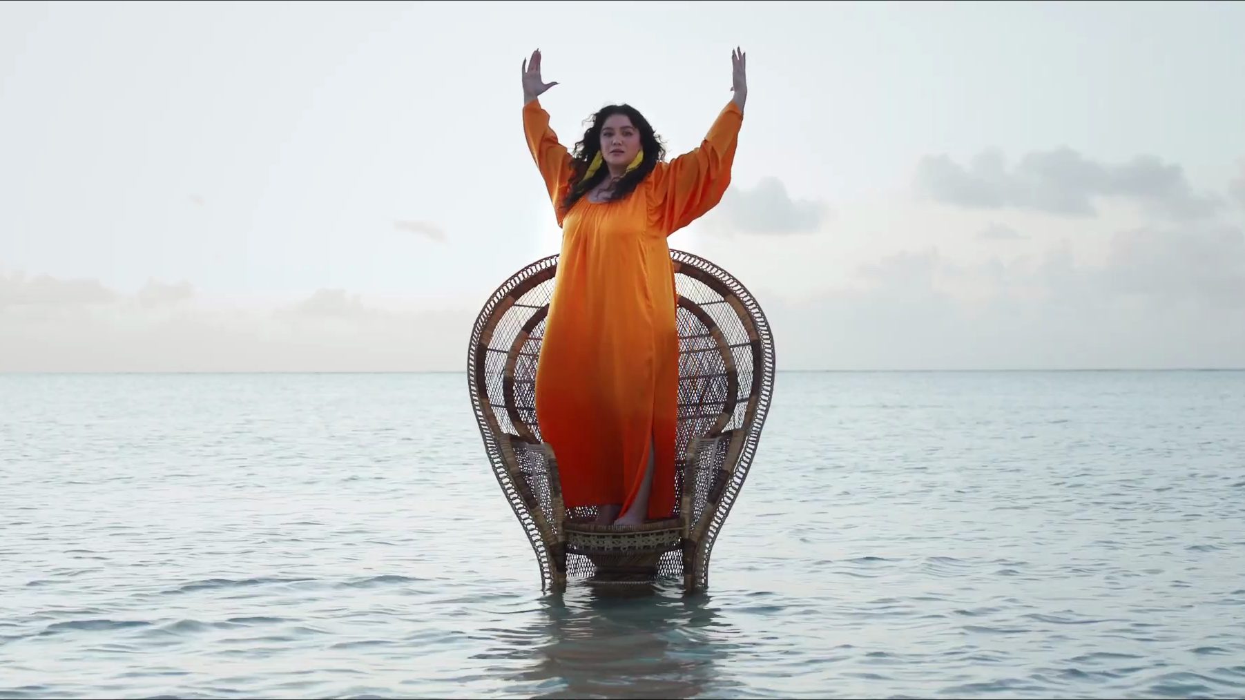 A woman in an orange dress stands with arms raised on a rattan chair in the sea